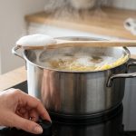 A hyper-realistic close-up of a stainless steel pot of pasta boiling vigorously on a spotless induction stovetop. Dense white foam rises rapidly, nearly overflowing, but is effectively contained below a light-colored wooden spoon balanced across the pot's rim. A hand gently turns a sleek control knob, managing the heat. Gentle steam rises, illuminated by bright natural light, with a blurred, minimalist kitchen in the background.