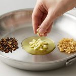 A top-down, close-up, photorealistic image of a pristine, brushed stainless steel skillet on a brightly lit, light kitchen counter. Inside the skillet, three distinct piles of minced garlic illustrate different cooking stages: On one side, a small pile of scorched, dark brown to black, shriveled garlic. In the center, a human hand gently drops bright white, fresh minced garlic into a shimmering pool of clear olive oil. On the opposite side, a pile of perfectly cooked, translucent golden-brown minced garlic. The background is a subtly blurred modern kitchen.