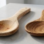 A photorealistic studio still life shows two wooden cooking spoons side-by-side on a clean counter. On the left, a worn, light-colored wooden spoon with an open grain is fuzzy, splintered, warped, and cracked, with deep, dark food stains absorbed into its coarse surface. In stark contrast, a pristine, premium spoon on the right, made from rich, dark, closed-grain wood (like olive wood), features an exceptionally smooth, polished, and unstained surface with a subtle sheen, highlighting its tight grain and perfect form. Both rest on a softly blurred, minimalist counter under bright natural light, emphasizing the stark visual contrast between worn utility and premium craftsmanship.