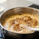 A close-up of deeply browned butter in a gleaming stainless steel saucepan, bubbling with golden-amber liquid and abundant, dark, intensely caramelized milk solids. A whisk rests on the pan's edge, with dry milk in the soft, golden-lit background.
