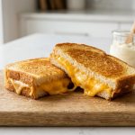 A hyper-realistic close-up of a flawlessly golden-brown grilled cheese sandwich, cut diagonally, with luscious, molten cheese oozing from the center. The halves rest on a rustic cutting board in a brightly lit, modern kitchen, with a soft-focused jar of mayonnaise in the background.