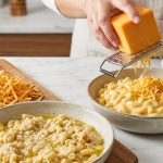 A photorealistic culinary scene dramatically contrasts two mac and cheese dishes. In the foreground, a bowl of mac and cheese has a lumpy, separated, dull orange sauce with oil visible, next to a pile of dry pre-shredded cheddar. In the background, skilled hands grate fresh cheddar over a second bowl, which holds a perfectly smooth, glossy, and rich golden cheese sauce.
