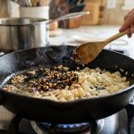 A close-up of a cast iron or stainless steel pan on a modern stovetop shows unevenly cooked aromatics. Dark brown, almost burnt minced garlic pieces are visible with faint smoke rising, contrasting sharply with the surrounding largely translucent and undercooked white onions. A home cook's hand, holding a wooden spoon, hovers above the pan, conveying slight concern in a clean, warmly lit kitchen.
