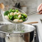 A photorealistic close-up shows a steady hand using a gleaming, wide-mesh stainless steel spider skimmer to gracefully lift vibrant green blanched broccoli florets and plump, intact white ricotta dumplings. Water actively drains and shimmers through the mesh as they are lifted from a steaming stainless steel pot. The modern kitchen scene is clean and brightly lit.