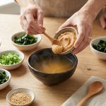 An overhead shot shows elegant hands gently scooping light brown miso paste into a bowl of clear, subtly steaming dashi broth. Around it, an array of small, clean ceramic bowls hold vibrant miso soup toppings: finely diced tofu, bright green scallions, deep green wakame, sliced shiitake mushrooms, and golden sesame seeds. The scene is set on a light wooden table, bathed in soft, diffused natural light, creating a clean, wholesome, and inviting atmosphere.