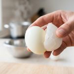 A close-up, photorealistic shot of a hand effortlessly peeling a perfectly hard-boiled egg, revealing a smooth, unmarred white surface. The shell separates cleanly, with a soft, minimalist kitchen background.