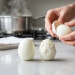 A high-resolution, ultra-photorealistic image captures a clean kitchen countertop with two hard-boiled eggs: one perfectly peeled and smooth, the other damaged with torn white and clinging shell. In the mid-ground, hands expertly peel a third egg cleanly. A steaming pot and tea towel in the soft-focus background hint at cooking. The bright, detailed scene conveys a satisfying and precise culinary moment.