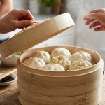 A photorealistic image of adult hands gently lifting the lid of a multi-tiered bamboo steamer, revealing a mix of perfectly plump, fluffy white bao buns and some subtly wrinkled ones. Delicate steam rises, illuminated by bright natural light in a clean kitchen with a softly blurred background, suggesting a home-cooked meal.