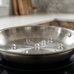 A photorealistic close-up shows an empty, gleaming stainless steel skillet on a stovetop, with spherical water droplets rapidly skittering and dancing across its superheated surface, vividly illustrating the Leidenfrost effect in a clean, modern kitchen.