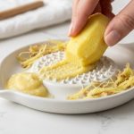 A close-up, photorealistic image of clean fingers grating fresh ginger on a white ceramic oroshigane grater. Smooth ginger puree accumulates on the grater, with tough fibers neatly separated around the edge, all on a bright, clean kitchen counter.