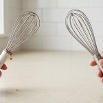 A photorealistic close-up on a clean, light kitchen countertop shows two whisks side-by-side. On the left, a hand gently tilts a used whisk with a hollow handle, revealing a rusty water droplet seeping from a visible seam and staining the handle. On the right, another hand confidently holds up a pristine, seamless, solid brushed stainless steel whisk, highlighting its hygienic and durable construction.