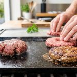 A photorealistic close-up shows burger patties cooking on a hot griddle, demonstrating a home cooking technique. On the left, a raw patty swells into a spherical shape. On the right, a dimpled raw patty and a perfectly flat, golden-brown cooked burger patty illustrate the successful technique, with home cook hands tending to them.