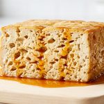 A close-up, hyperrealistic photo of a cut tofu block revealing a highly porous, sponge-like interior, deeply saturated and glistening with a vibrant amber marinade. A small pool of marinade gathers at its base. Presented on a clean, light-colored surface with a softly blurred kitchen background.