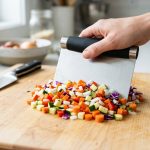 A close-up of a hand expertly using a sleek stainless steel bench scraper to neatly gather a vibrant pile of freshly chopped colorful vegetables on a clean wooden cutting board in a brightly lit kitchen. A gleaming chef's knife rests in the soft-focus background.