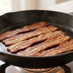 A photorealistic close-up of several perfectly cooked, golden-brown, crispy streaky bacon strips glistening with rendered fat in a clean, seasoned cast iron skillet, resting on a gas stovetop with a softly blurred, warm kitchen background, conveying culinary perfection.