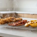 A sturdy stainless steel cooling rack holds perfectly crispy, golden-brown fried chicken, bacon, and roasted potato wedges on a clean kitchen counter, with subtle steam rising from underneath the food, emphasizing its freshness and excellent airflow.