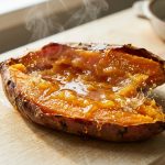 A perfectly roasted sweet potato, split open lengthwise, reveals an incredibly gooey, syrupy, translucent amber interior glistening intensely with natural sugars. Delicate wisps of steam rise from its warm surface. Subtle shimmering golden particles around the edges visually represent the potato's starches transforming into flowing sugar. The ultra-photorealistic close-up shot features a shallow depth of field, blurring a light wooden background.