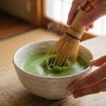 A close-up, ultra-photorealistic image of a human hand using a bamboo whisk to froth vibrant jade-green matcha tea in a minimalist ceramic bowl. A perfect, creamy foam forms on the tea's surface, illuminated by soft natural light against a serene, blurred background, conveying a sense of calm and mindful preparation.