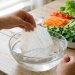 Close-up of hands gently submerging a dry rice paper wrapper into a clear glass bowl of water, which is softening and becoming translucent. In the softly blurred background, vibrant spring roll ingredients like julienned carrots, lettuce, and cooked shrimp are artfully arranged on a light wooden table. The scene is bright, clean, and well-lit by natural light.