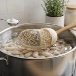 A photorealistic close-up shows dried beans simmering in a sleek stainless steel pot in a clean, minimalist kitchen. One bean reveals incredibly detailed, rigid pectin structures resistant to softening, while a neighboring bean shows visibly loosened, hydrated pectin. A spoon holding baking soda hovers, creating a ripple in the water, symbolizing alkalinity. On a pristine counter, a sliced tomato and lemon are artfully arranged, signifying acidic ingredients not yet added. The image conveys precise scientific cooking in a high-definition, molecular detail.