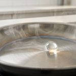 A perfectly spherical, glowing water droplet rapidly skitters across an ultra-hot, gleaming stainless steel frying pan, enveloped by a thin cushion of steam. The pan shows subtle heat discoloration and mirror-like reflections, with a softly blurred modern kitchen in the background. Ultra photorealistic.