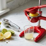 A photorealistic still life on a pristine counter shows a gleaming red citrus press expertly squeezing a lemon, with a clear juice stream flowing into a glass and a perfectly spent lemon half nearby. This efficiency contrasts sharply with scattered, partially squeezed fruit, rogue seeds, and worn, discolored plastic and dull aluminum juicers, all bathed in bright natural light.