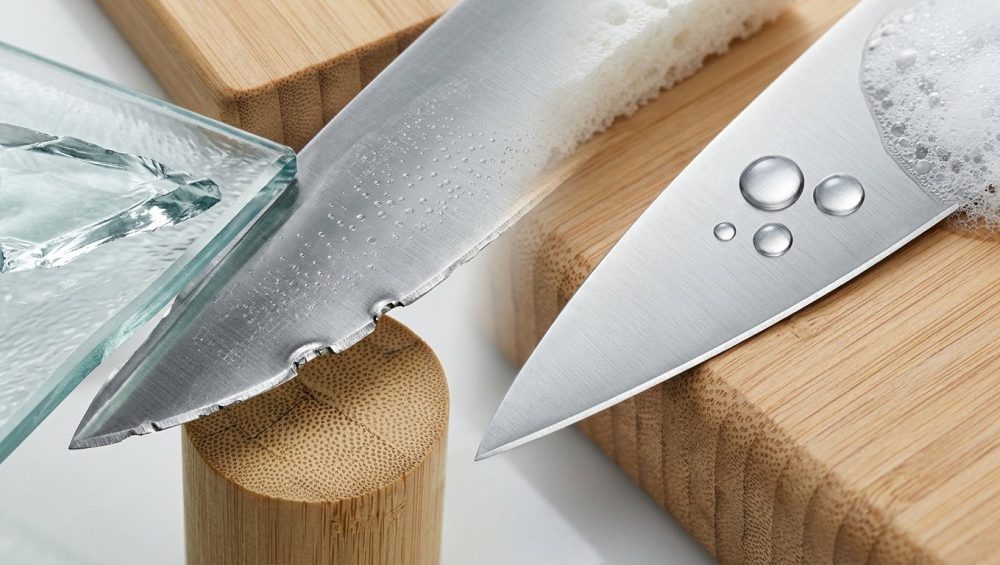 A detailed, photorealistic macro image of a kitchen knife blade, visually comparing a damaged edge with a perfectly preserved one. The damaged section shows a rolled and chipped edge, next to sharp glass shards and dense bamboo fibers, with subtle etching from harsh cleaning. In contrast, the pristine section rests gently on a soft wood cutting board, with clear water droplets and fine soap suds illustrating proper hand-washing and care. The bright, analytical lighting emphasizes textures and metallic sheen, in a clean, scientific style.