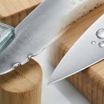 A detailed, photorealistic macro image of a kitchen knife blade, visually comparing a damaged edge with a perfectly preserved one. The damaged section shows a rolled and chipped edge, next to sharp glass shards and dense bamboo fibers, with subtle etching from harsh cleaning. In contrast, the pristine section rests gently on a soft wood cutting board, with clear water droplets and fine soap suds illustrating proper hand-washing and care. The bright, analytical lighting emphasizes textures and metallic sheen, in a clean, scientific style.