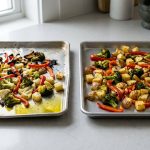 A photorealistic studio shot shows two baking sheets side-by-side on a clean kitchen counter. On the left, a warped, flimsy aluminum pan holds unevenly cooked vegetables – some charred, some pale – and a pool of olive oil. On the right, a pristine, heavy-gauge steel pan displays uniformly golden-brown, perfectly roasted vegetables, highlighting the stark difference in cooking performance and pan quality.