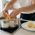 A bright, photorealistic image shows a home cook's hands lowering raw potato chunks into a stainless steel pot of cold water on an induction stove. Beside the pot, a halved, perfectly cooked potato chunk reveals its uniformly tender, fluffy texture, illustrating the "cold start" cooking method for consistent results.