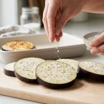 Close-up of hands sprinkling coarse sea salt on raw eggplant slices, which are visibly beading with moisture. A golden, cooked eggplant slice rests in the soft-focus background.