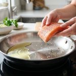 A high-angle, photorealistic close-up of a gleaming stainless steel frying pan on a modern stovetop. Water droplets dance from the Leidenfrost effect next to spreading cooking oil, while capable hands hold raw food, poised to cook, in a clean, minimalist kitchen.