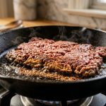 Close-up of ground beef searing in a hot cast iron pan, featuring a deep golden-brown, caramelized crust on the bottom and a vibrant reddish-pink top, with subtle steam rising.