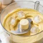 Ultra-photorealistic close-up of a modern food processor actively blending silky, pale hummus, with several whole, crystalline ice cubes visible in the swirling mixture. The setting is a bright, clean kitchen.
