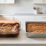 A photorealistic studio comparison on a sleek countertop shows two trays of brownies. The left tray, in a clear glass dish, has noticeably dark, crisp edges and a light, gooey center, with a subtle orange heat haze. The right tray, in a dark metal pan, is uniformly golden-brown and perfectly baked from edge to center, illustrating ideal, even results.
