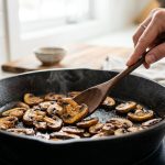 Photorealistic close-up of perfectly caramelized, crispy cremini mushrooms searing in a dry cast iron skillet, gently stirred by a hand with a wooden spoon against a blurred, bright modern kitchen background.
