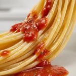 A macro, photorealistic studio shot of golden spaghetti coated in glistening olive oil. Vibrant red tomato sauce, flecked with herbs, is being drizzled onto the pasta but visibly beads up and slides off, failing to cling to the slick surface. Small droplets gather, leaving much of the pasta bare, while the sauce pools below, clearly illustrating the lack of adhesion. Sharp focus highlights this interaction against a clean, blurred background.