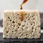 Close-up studio shot of a thawed tofu cross-section, revealing its intricate, highly porous, sponge-like honeycomb structure. A glistening amber marinade drop hovers just above, showing its readiness to absorb flavor.