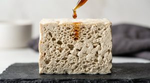 Close-up studio shot of a thawed tofu cross-section, revealing its intricate, highly porous, sponge-like honeycomb structure. A glistening amber marinade drop hovers just above, showing its readiness to absorb flavor.