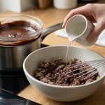 A photorealistic close-up shows a hand pouring hot liquid into a bowl of dull, grainy, seized chocolate paste, which a whisk is starting to stir. In the background, a smaller bowl holds perfectly smooth, glossy dark chocolate over a double-boiler with rising steam. The clean, brightly lit scene contrasts the textures of the two chocolates, illustrating a culinary solution.