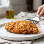 A perfectly pan-fried, golden-brown schnitzel or fish fillet rests on a white plate. Its uniformly crispy breading is completely intact, showing no signs of peeling. A wisp of steam rises from the hot food, with a home cook's hands holding tongs visible in the soft-focus background of a clean kitchen.