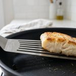 A close-up shows a sleek, ultra-thin metal fish spatula precisely sliding completely under a perfectly pan-seared, flaky fish fillet in a non-stick pan, with a softly blurred minimalist kitchen background.