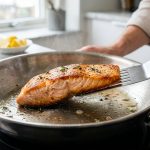 A photorealistic close-up of a salmon fillet searing in a gleaming stainless steel skillet. A spatula lifts a corner, revealing a flawless, crispy, golden-brown Maillard reaction crust. The exposed flesh looks tender and moist, with no residue on the pan, all captured with bright, professional food photography.