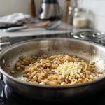 A close-up of a professional-grade stainless steel sauté pan on a sleek stovetop. Inside, finely diced yellow onions are actively sautéing to a translucent, golden-brown, while freshly minced, pale ivory garlic has just been introduced alongside them, clearly less cooked. Subtle steam rises under soft, natural daylight in a softly blurred, minimalist kitchen.