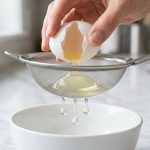 An ultra-photorealistic close-up of a hand gently cracking a raw egg over a fine-mesh strainer. Thin, watery egg white drips in clear drops into a white bowl below, while the thick, gelatinous albumen remains held within the mesh, set against a clean, bright kitchen counter.