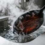 A faintly glowing red cast iron skillet is plunged into cold water in a modern kitchen sink. A dramatic plume of thick white steam erupts as the skillet's base visibly warps and buckles from the severe thermal shock. The close-up shot highlights the powerful, destructive reaction in a pristine, high-contrast setting.