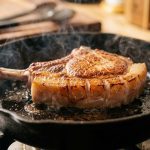 An ultra-photorealistic close-up of a thick-cut, bone-in pork chop actively searing in a hot cast iron skillet. The meat displays a flawless, uniform, deep brown Maillard crust. Its scored fat cap is glistening, rendered, and gently bubbling, with subtle steam rising, conveying intense heat and successful cooking.