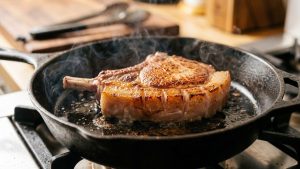 An ultra-photorealistic close-up of a thick-cut, bone-in pork chop actively searing in a hot cast iron skillet. The meat displays a flawless, uniform, deep brown Maillard crust. Its scored fat cap is glistening, rendered, and gently bubbling, with subtle steam rising, conveying intense heat and successful cooking.
