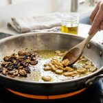 A highly detailed, photorealistic close-up of a sauté pan on a stovetop, illustrating a stark contrast: burnt, dark garlic on the left side, and perfectly golden, translucent garlic gently sautéing in olive oil on the right, being stirred by hands.