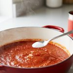 A photorealistic close-up of vibrant, deep red tomato pasta sauce gently simmering in a modern enameled pot. A silver spoon, perfectly centered, suspends a tiny pinch of white baking soda just above the bubbling surface, from which subtle steam rises. In the softly blurred background, an unbranded can of whole peeled tomatoes hints at the ingredients on a clean counter. The scene evokes practical culinary science.